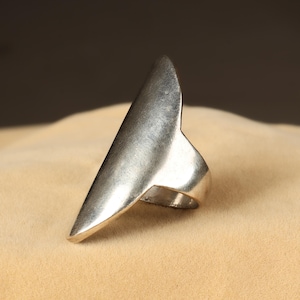 May include: A silver ring with a distinctive, elongated design. The ring has a smooth, metallic finish and a pointed top. The ring's design extends up the finger, creating a bold and modern look. A statement piece of jewellery.