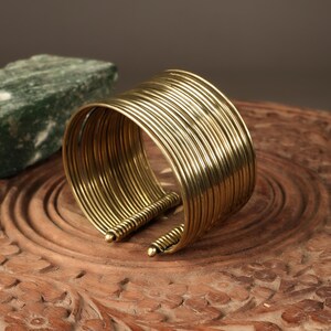 Op de afbeelding: Een brede, open manchetarmband gemaakt van meerdere dunne, goudkleurige metalen banden. De armband heeft een afgeronde vorm en wordt gepresenteerd op een gesneden houten oppervlak. De manchet is ongeveer 5 cm breed.