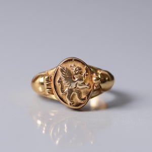 Könnte beinhalten: Ein goldener Ring mit ovaler Fassung, die ein detailliertes Greifen-Design zeigt. Der Ring hat einen abgerundeten Schaft und eine polierte Oberfläche, die das Licht reflektiert. Der Greif ist mit Flügeln und einem Löwenkörper dargestellt.