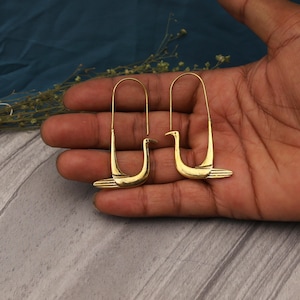 May include: A pair of gold-toned metal earrings featuring a stylized bird silhouette. The earrings have a long, U-shaped wire hook that connects to the base of the bird figure, which includes a pointed beak and horizontal lines on the tail section.