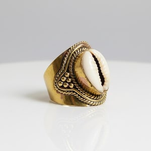 May include: A brass ring featuring a white cowrie shell. The ring has a wide band with decorative rope and dot detailing around the shell. The ring is set against a white background.