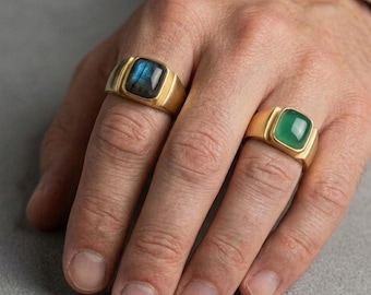 May include: Two gold signet rings with square gemstones. One ring features a blue-gray stone, the other a green stone. The rings are on a hand, showcasing the jewelry's design and color.