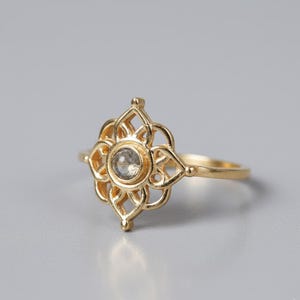 Pode incluir: Um anel dourado com um design floral. O anel apresenta uma pedra central, redonda e transparente, rodeada por uma flor vazada de oito pétalas. A banda é fina e lisa, e o anel está sobre um fundo cinza claro.