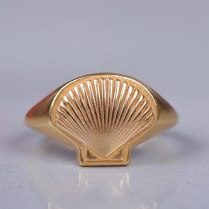 Puede incluir: Un anillo dorado con diseño de concha. El anillo presenta un patrón de concha detallado y en relieve sobre una superficie ovalada y plana. La banda es lisa y redondeada, y el anillo está sobre un fondo claro.