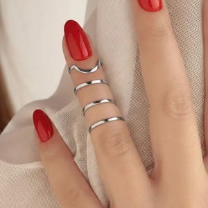 Puede incluir: Una mano con esmalte de uñas rojo luce anillos plateados. Los anillos incluyen un diseño ondulado y varias bandas finas, creando un efecto en capas. Los anillos son minimalistas y modernos, adecuados para el uso diario.