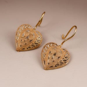 Puede incluir: Un par de pendientes dorados en forma de corazón. Los pendientes presentan un diseño calado con detalles intrincados, incluyendo patrones en forma de hojas y acentos circulares. Los ganchos para las orejas son curvos y están unidos a la parte superior.