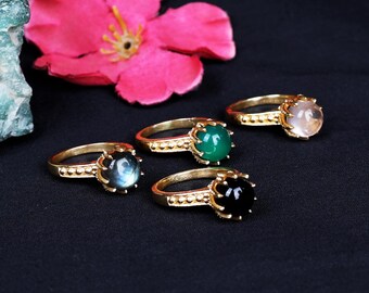 May include: A collection of gold-toned rings, each featuring a different colored gemstone: blue, green, black, and clear. The rings are arranged on a dark surface, with a pink flower in the background.