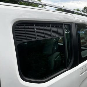 May include: A white van with a black window vent and tinted windows. The side of the vehicle is visible, showcasing the window design and the vehicle's overall shape. The roof has a black luggage rack.
