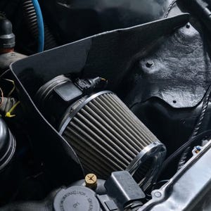 Könnte beinhalten: Nahaufnahme eines Motorraums mit einem zylindrischen Luftfilter. Der Filter ist silber und grau, befestigt mit einer Metallklemme. Ein schwarzes, rechteckiges Gehäuse umgibt den Filter. Verschiedene Schläuche und Komponenten sind sichtbar.