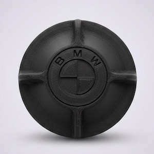 Könnte beinhalten: Schwarzes, kreisförmiges Autoteil mit dem BMW-Logo in der Mitte. Das Teil hat eine strukturierte Oberfläche und ein kreuzförmiges Design. Das BMW-Logo ist weiß und von den Buchstaben "B", "M" und "W" umgeben.