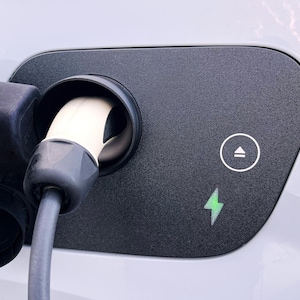 Könnte beinhalten: Nahaufnahme einer Ladebuchse für Elektrofahrzeuge. Die schwarze Anschlussabdeckung hat einen weißen Kreis mit einem Linien- und Pfeilsymbol und ein grünes Blitzsymbol. Ein weißes Ladekabel ist eingesteckt.