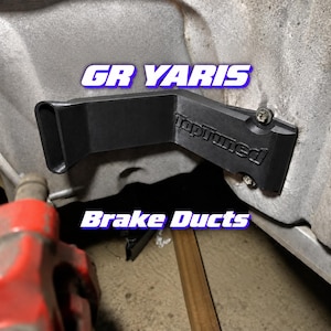 Könnte beinhalten: Ein schwarzes, L-förmiges Bremsluftführungsteil für einen GR Yaris mit dem eingeprägten Schriftzug TopTuned, befestigt mit zwei Schrauben. Das Bauteil ist an einer Metalloberfläche angebracht, überlagert mit dem Text GR YARIS und Brake Ducts.