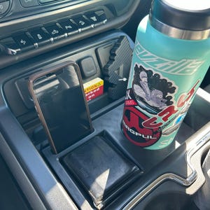 Puede incluir: Consola central de coche con teléfono, cartera y botella de agua. El teléfono está en una funda marrón. La botella de agua es turquesa con pegatinas. La cartera es negra. La consola es gris.