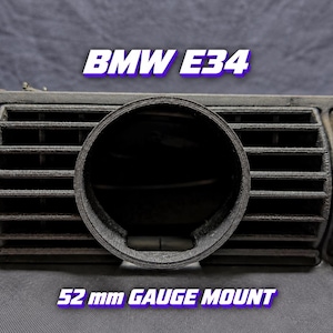 Op de afbeelding: Een zwarte 3D-geprinte ronde metermount ontworpen voor een BMW E34 dashboard ventilatierooster. De mount heeft een opening van 52 mm in een horizontaal lamellenrooster. De tekst BMW E34 en 52 mm GAUGE MOUNT is zichtbaar op de afbeelding.