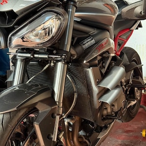 May include: A grey and black motorcycle with the text "Street Triple" visible. The motorcycle features a headlight, front suspension, and a red frame. The tyres are black.