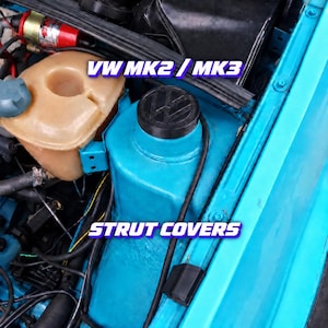 Könnte beinhalten: Eine schwarze, runde Federbeindomkappe mit geprägtem VW-Logo, montiert auf einem hellblau lackierten Federbeindom im Motorraum. Der Text VW MK2 / MK3 STRUT COVERS ist in weißer und violetter Schrift über das Bild gelegt.