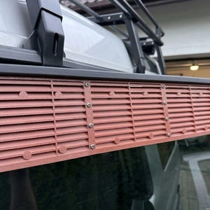 Peut inclure: Un évent à persiennes brun-rouge installé sur la fenêtre d'un véhicule. L'évent rectangulaire comporte des lattes horizontales et est fixé avec des vis argentées. Le véhicule est équipé d'un porte-bagages noir.