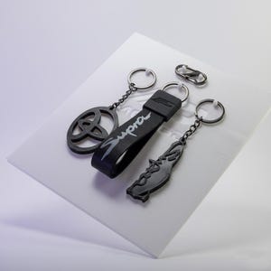 Può includere: Una collezione di portachiavi neri e un moschettone argentato. I portachiavi presentano vari design, tra cui un logo, la parola "Supra" e una forma stilizzata. I portachiavi sono esposti su una superficie bianca.