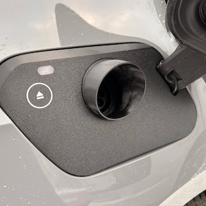 May include: Close-up of a grey car's charging port. The port has a black, cylindrical opening and a textured, dark grey cover with a white indicator light and a circular button with an upward arrow symbol.