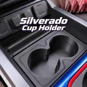 Puede incluir: Un portavasos doble de plástico negro para la consola central de un vehículo con el texto Silverado Cup Holder impreso en blanco. El inserto cuenta con dos aberturas circulares y está colocado dentro de un área de consola gris oscuro con tres cubiertas.