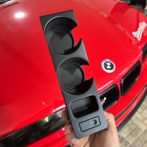 Può includere: Un inserto per console centrale automobilistica in plastica nera opaca dotato di due portabicchieri circolari e un vano portaoggetti rettangolare con un piccolo pulsante quadrato alla base, progettato per la configurazione del cruscotto.