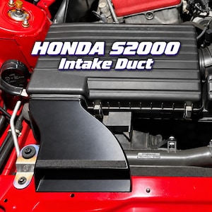 May include: A black Honda S2000 Intake Duct installed within a red engine bay. The component features a smooth, matte black finish and is positioned next to a black air filter housing. The text HONDA S2000 Intake Duct is overlaid in white.