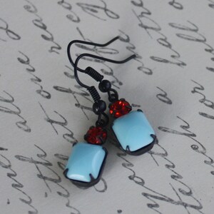 Op de afbeelding: Een paar oorhangers met blauwe en rode glazen stenen. De oorbellen hebben zwarte metalen haakjes en zijn versierd met kleine zwarte kralen.