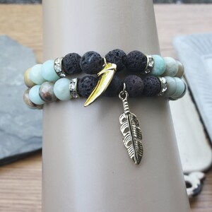 May include: Two layered bracelets with black lava rock beads, light blue and brown agate beads, and gold accents. One charm is a gold feather and the other is a gold tooth.