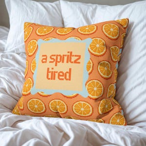 Könnte beinhalten: Quadratisches orangefarbenes Kissen mit einem sich wiederholenden Muster aus Orangenscheiben. In der Mitte befindet sich ein hellorangefarbenes Quadrat mit den Worten "a spritz tired" in orangefarbener Pixel-Schrift, umrahmt von einem hellblauen Rand. Das Kissen liegt auf einem weißen Bett.