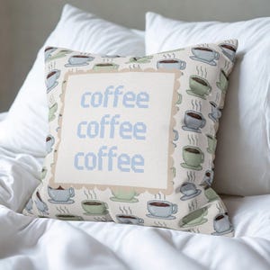 May include: Square decorative pillow with a repeating pattern of coffee cups in shades of blue, green, and brown on a cream background. The center features the word "coffee" repeated three times in blue pixelated font.