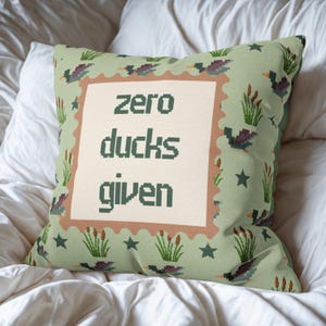 May include: Square decorative pillow with a light green background and a repeating pattern of ducks, cattails, and stars. The center features the phrase "zero ducks given" in dark green block letters within a cream-colored frame.