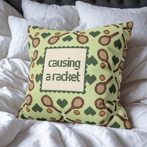 May include: A square decorative pillow with a light green background featuring a pattern of tennis rackets, tennis balls, and green hearts. The center of the pillow has the text "causing a racket" in a pixelated font.