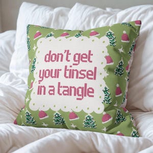 Peut inclure: Un coussin carré décoratif avec un fond vert orné de motifs de bonnets de Père Noël roses et de sapins de Noël pixélisés. Le coussin porte l'inscription rose : "don't get your tinsel in a tangle".