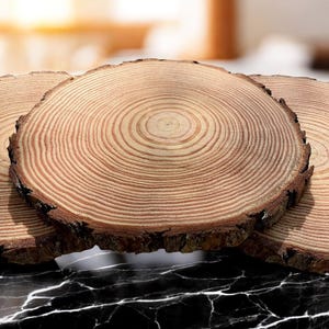 Könnte beinhalten: Drei runde Untersetzer aus Holz mit sichtbaren Jahresringen und einem natürlichen Rindenrand. Die Untersetzer sind hellbraun mit dunkleren braunen Ringen, die die Maserung des Holzes zeigen. Sie sind auf einer schwarz-weißen Marmoroberfläche angeordnet.