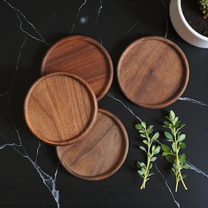 May include: Four round wooden coasters with a raised edge, showcasing a rich brown wood grain. The coasters are arranged on a black marble surface, with sprigs of green foliage and a white potted plant in the background. A simple, natural aesthetic.
