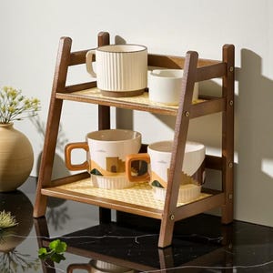 Puede incluir: Una estantería de madera de dos niveles con base tejida sostiene varias tazas de cerámica. Las tazas son de color crema con detalles marrones y presentan varios diseños. La estantería es de madera oscura y tiene una estructura similar a una escalera.