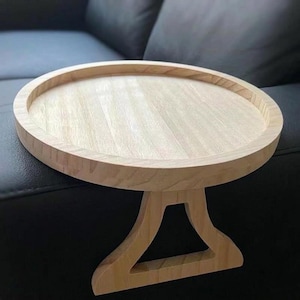 Puede incluir: Pequeña mesa auxiliar redonda de madera clara con borde elevado. Tiene una sola pata decorativa. Diseñada para colocarse junto a un sofá o sillón.