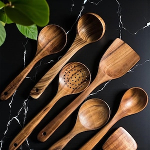 Peut inclure: Ensemble d'ustensiles de cuisine en bois, comprenant des cuillères, une spatule et une cuillère ajourée. Les ustensiles sont en bois brun clair avec des motifs de grain visibles. Les articles sont disposés sur une surface noire veinée de blanc.
