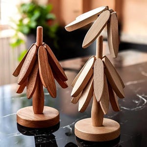 Puede incluir: Dos esculturas decorativas de madera. Cada escultura tiene una base redonda, un tallo central y numerosas piezas de madera en forma de hoja que irradian hacia afuera. La madera tiene un tono cálido y natural. Las esculturas se exhiben sobre una superficie negra.