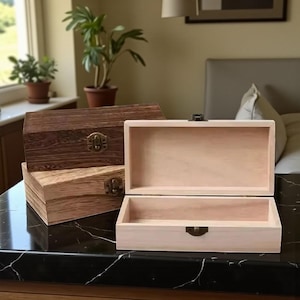 May include: Three wooden boxes with hinged lids and metal clasps. One box is open, revealing the natural wood interior. The other two boxes are stacked, showcasing the different wood grains and finishes. The boxes are on a black marble surface.