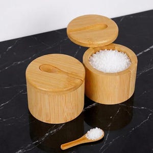 Może przedstawiać: Dwie bambusowe solniczki z pokrywkami i małymi drewnianymi łyżeczkami. Jedna solniczka jest otwarta, wypełniona białą solą. Druga jest zamknięta. Solniczki stoją na czarnej marmurowej powierzchni.