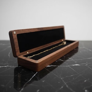 Puede incluir: Caja de joyería de madera abierta con interior de terciopelo negro. Un collar de oro descansa dentro de la caja. La caja es de madera oscura y tiene un cierre magnético. La caja está sobre una superficie de mármol negro.