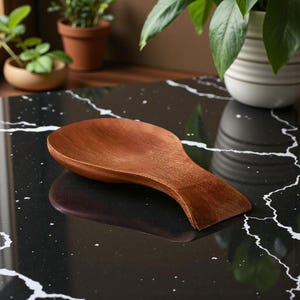 May include: A wooden spoon rest with a curved handle. The spoon rest is made of a dark brown wood and is resting on a black surface with white veining. The spoon rest is designed to hold a spoon.