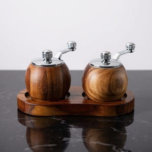 May include: A set of wooden salt and pepper grinders. Each grinder has a silver top and handle. They sit on a wooden tray. The wood has a natural grain. The set is designed for the dining table.