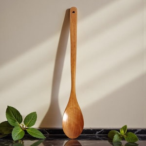 May include: A long, wooden spoon with a rounded bowl and a handle with a small hole. The spoon is made of light brown wood and is set against a neutral background. The spoon is approximately 12 inches long.