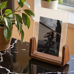 May include: A wooden photo frame with a clear acrylic front displays a photograph. The frame has a warm, natural wood finish and a unique curved design. The photo shows a woman in a floral dress. The frame sits on a black and white marble surface.
