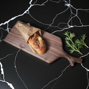 Può includere: Un tagliere in legno con manico, che tiene una pagnotta di pane avvolta in carta marrone. Il pane è marrone dorato con semi di sesamo. Erbe verdi fresche sono poste di lato.