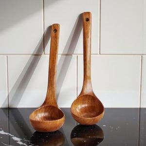 May include: Two wooden ladles with long handles and rounded bowls. The ladles are made of a light brown wood and have a hole at the end of the handle for hanging. The ladles are sitting on a black surface.
