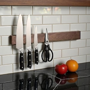 May include: A wooden magnetic knife holder with four knives and a pair of scissors attached. The knives have black handles and silver blades. A red tomato and orange sit on the countertop below the knives.