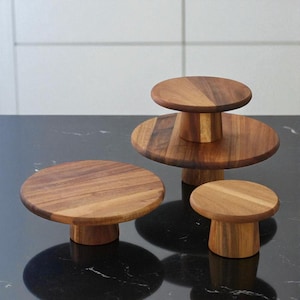 May include: A collection of three wooden cake stands. The stands are crafted from warm-toned wood, showcasing a natural grain. Each stand features a circular top and a short, cylindrical base. They are presented on a dark surface.
